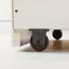 A close-up view of the bottom of a white bed frame, showing a black caster wheel attached to the wooden frame. Produktbild-Vorschau