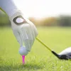 A close-up shot of a hand in a white golf glove placing a white golf ball on a pink tee on a golf course. Produktbild-Vorschau