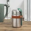 A stainless steel food flask with a brown leather strap and a green spoon attached, placed on a wooden surface next to a green mug and some books. Náhled obrázku produktu