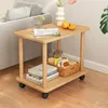 A small wooden side table with two shelves is placed next to a sofa, holding various items like books, a basket of snacks, and fruit. Produktbild-Vorschau