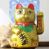 A golden Maneki Neko cat figurine sits on a surface with a blurred blue and white patterned background and a shell-like object to the left. Produktbild-Vorschau