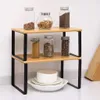 A two-tier shelf organizer holding various jars of spices and ingredients, with a stack of white bowls and plates on the bottom shelf. Produktbild-Vorschau