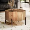 A round wooden coffee table with a vase of dried flowers on top, placed on a rug in a living room. Produktbild-Vorschau