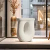 A white, abstract-shaped side table is displayed on a light gray surface in front of a window with a blurred city view. Náhled obrázku produktu
