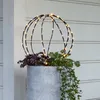 A decorative sphere with lights sits in a planter with plants. Produktbild-Vorschau