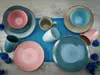 A collection of ceramic plates and mugs arranged on a wooden surface with a blue placemat. Náhľad obrázku produktu