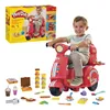 A young boy is riding a red Play-Doh pizza delivery scooter. Various Play-Doh accessories are scattered around the scooter. Produktbild-Vorschau