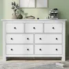 A white wooden dresser with seven drawers is shown in a room with a light green wall and a rug on the floor. Decorative items are placed on top of the dresser. Produktbild-Vorschau