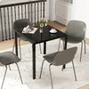 A square black glass-top dining table with four gray chairs around it, set on a white rug in a dining area. Produktbild-Vorschau