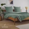 A neatly made bed with green bedding and pillows, a plant in a basket, and a vase with dried flowers in the background. Voir les images du produit