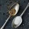 Two spoons, one filled with salt and the other with pepper, are placed on a dark surface. Produktbild-Vorschau