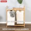 A bamboo laundry sorter with two fabric bags, placed on a wooden floor against a light gray wall. There are folded towels on the top shelf and a plant in a vase in the background. Podgląd zdjęcia produktu