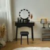 A black vanity table with a lighted mirror and a stool, set against a white wall with a window and curtains on the left, and a gray side table with a lamp and clock on the right. Voir les images du produit