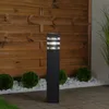 A black bollard light with clear glass panels is placed in a garden setting, with a wooden fence in the background and decorative white stones and small green bushes in the foreground. Podgląd zdjęcia produktu