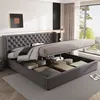 A bedroom with a large grey upholstered bed with its storage compartment open, revealing stored items. A large window with curtains is visible in the background. Náhled obrázku produktu