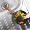 A hand is cleaning a dirty frying pan in a kitchen sink with a green sponge. Produktbild-Vorschau