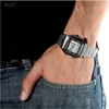 A person's arm wearing a silver digital watch with a black strap, with their hand in their jeans pocket. Produktbild-Vorschau