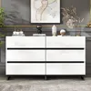 A white, high-gloss sideboard with six drawers is displayed in a room with gray walls and a framed abstract artwork above it. Decorative items are placed on top of the sideboard. Produktbild-Vorschau