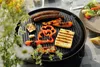 A close-up of a grill with food cooking on it, set outdoors on a table with flowers in the foreground. Podgląd zdjęcia produktu