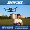 A man is flying a drone in a grassy field with buildings in the background. Produktbild-Vorschau