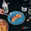 A hand is using tongs to place pasta onto a blue plate, with a bottle of wine and a bottle of olive oil in the background. Produktbild-Vorschau
