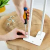 A person is using a screwdriver to attach a white hairpin leg to a wooden surface. Produktbild-Vorschau