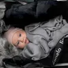 A baby is nestled in a stroller or bassinet, wrapped in a gray blanket and wearing a pink hat. Náhled obrázku produktu