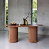 A modern dining room with a concrete wall and large windows, featuring an oval wooden dining table with two fluted pedestals. The table is set with a bowl of fruit, a striped cup, and two decorative vases with branches. Produktbild-Vorschau