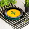 A close-up shot of a black bowl filled with yellow soup, garnished with a basil leaf, placed on a black and white striped placemat. Anteprima dell’immagine prodotto
