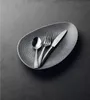 A set of cutlery (spoon, fork, and knife) is placed on a textured, oval-shaped dark gray plate, which is set on a dark gray surface. Produktbild-Vorschau