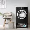 A black washing machine stand with a shelf is shown in a laundry room setting, next to a chair with pillows and a laundry basket. Náhled obrázku produktu