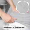 Hands stretching a light gray fitted sheet over a mattress, with a close-up inset showing the elasticated edge. Produktbild-Vorschau