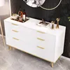 A white dresser with gold handles and legs, decorated with bottles, glasses, and decorative items, set against a black ribbed wall and a circular mirror. Podgląd zdjęcia produktu