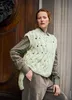 A woman wearing a light green knitted vest over a brown shirt and brown pants, leaning against a wooden door. Produktbild-Vorschau