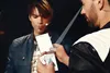 Two men are on a stage, one holding a card with geometric shapes and the other looking at it. Produktbild-Vorschau