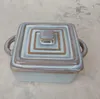 A square ceramic dish with a lid and handles sits on a textured surface. Náhled obrázku produktu
