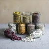 Several glass jars filled with various dry goods, such as pasta, beans, crackers, and grains, are arranged in a pyramid formation on a gray surface against a textured tan background. Podgląd zdjęcia produktu