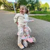 A young child is happily sitting on a pink and white balance scooter in a park. Podgląd zdjęcia produktu