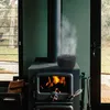 A cast iron humidifier sits on top of a wood-burning stove, emitting steam. The stove is in a room with a large window overlooking a natural landscape. Produktbild-Vorschau