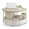 A round coffee table with a storage basket underneath, containing a pillow, a folded blanket, and books. A laptop and a small plant are on top of the table. Produktbild-Vorschau