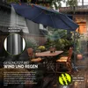 An outdoor patio scene with a large navy blue cantilever umbrella providing shade over a wooden table and chairs. It is raining, and leaves are falling from the umbrella. Náhled obrázku produktu