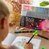 A child's hands are shown coloring in a book with markers from a set of RAGI dual tip marker pens. The marker set is displayed prominently in the background, along with some decorative dried flowers and candies. Produktbild-Vorschau