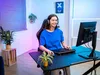 A young woman is playing a video game on a computer in a room with colorful lighting. Produktbild-Vorschau