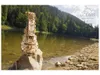 A wooden mechanical model of a hurdy-gurdy is placed on a rock by a lake, with a forest-covered mountain in the background. Produktbild-Vorschau