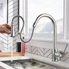 A hand is shown operating a modern kitchen faucet with a pull-down sprayer, with water flowing into a sink. The background features a window with a snowy landscape and a tiled backsplash. Náhľad obrázku produktu