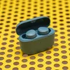 A pair of teal wireless earbuds in their open charging case, resting on a yellow perforated surface. Anteprima dell’immagine prodotto