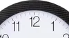 A close-up view of a white analog clock face with black numbers and markings, set within a black carbon fiber-patterned frame. Voir les images du produit