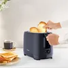 A dark gray toaster is on a white countertop, with two slices of bread inside, and another hand reaching for a slice. There is also a cup of coffee and a plate of toast in the foreground. Produktbild-Vorschau