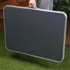 A person is holding a folded camping table outdoors on a grassy area. Produktbild-Vorschau