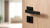 A close-up shot of a black door handle and keyhole on a wooden door. The background is blurred, showing a living room interior. Produktbild-Vorschau
